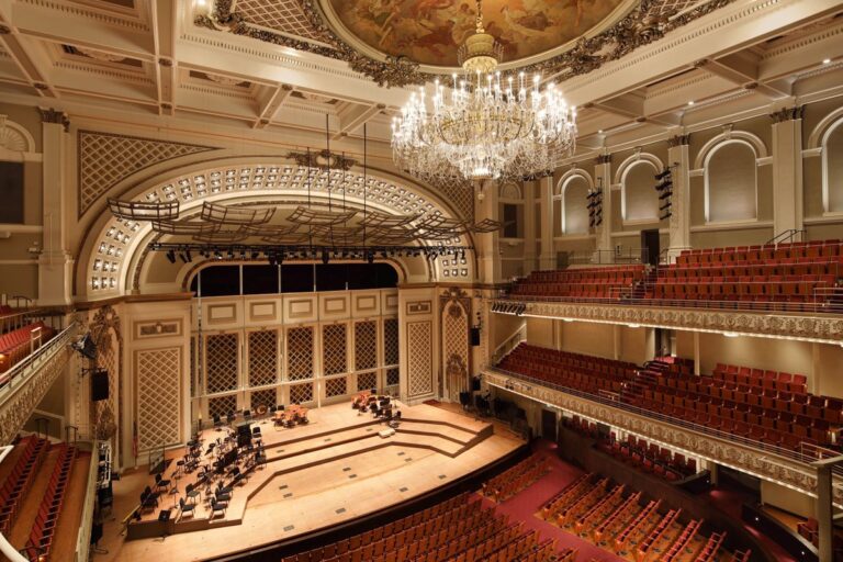 Image of Springer Auditorium from the audience with red chairs, crystal chandelier, ornate ceilings, and empty stage.