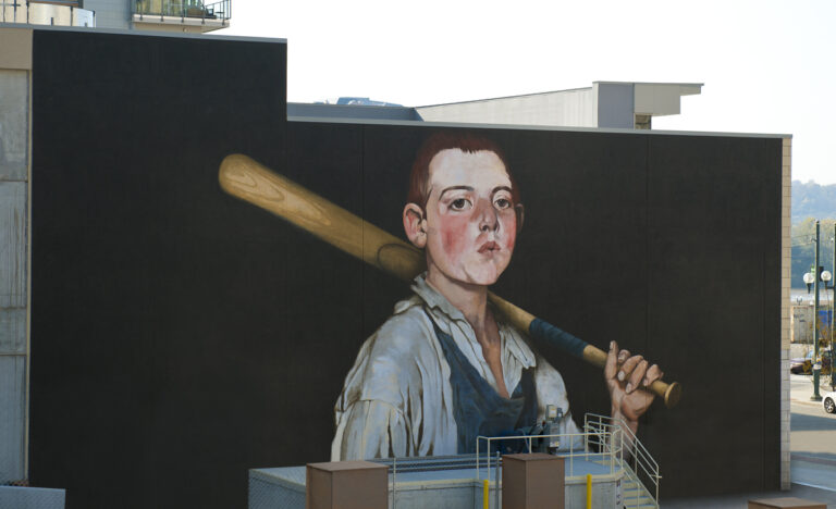 Close-up photo of “The Cobbler’s Apprentice Plays Ball” mural. A young boy wears blue overalls and whistles while holding a baseball bat over his shoulder.